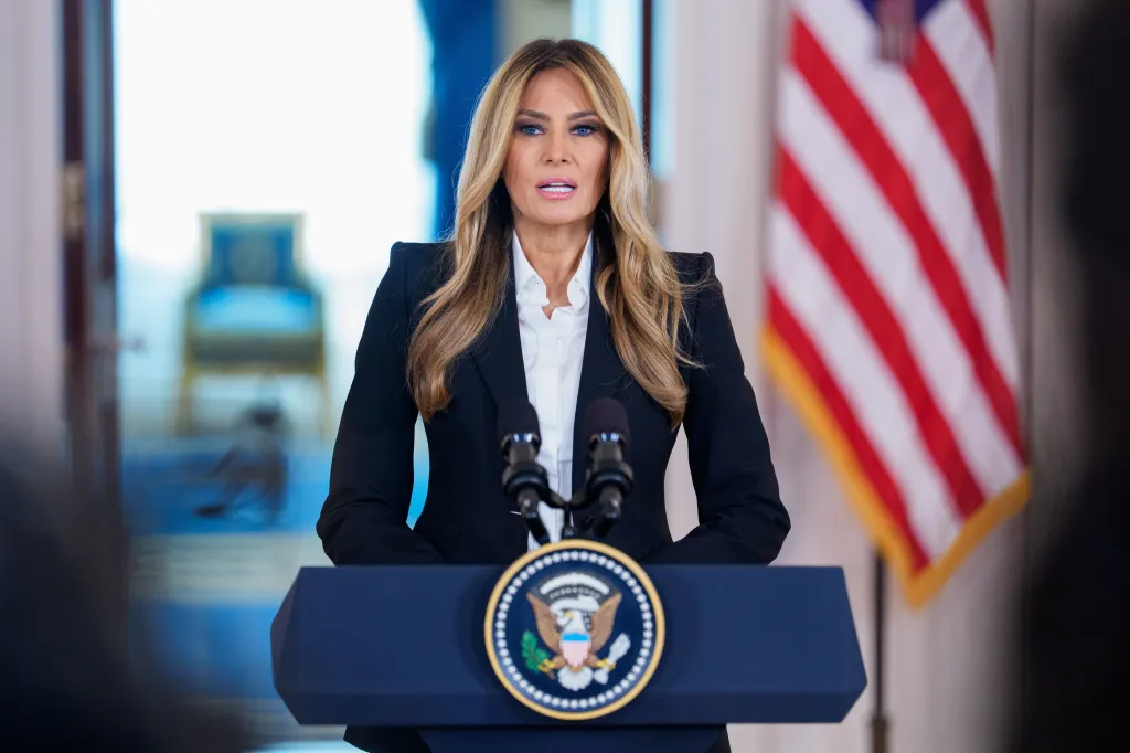Melania Trump speaking at a podium with two microphones, an American flag in the background.