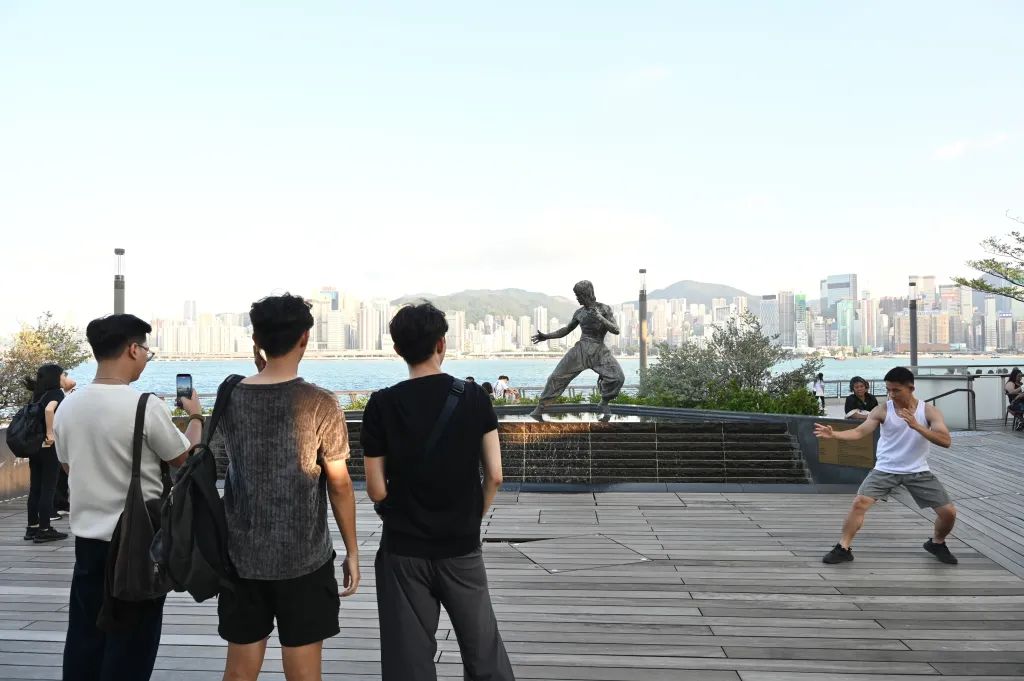 A man posing like Bruce Lee in front of the Bruce Lee statue in Hong Kong, with a cityscape and water in the background.