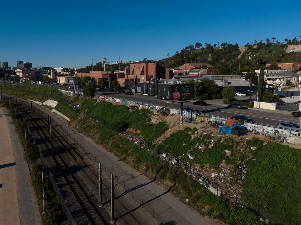 Homeless encampments and debris on a grassy hillside next to a railway and a road in Los Angeles.