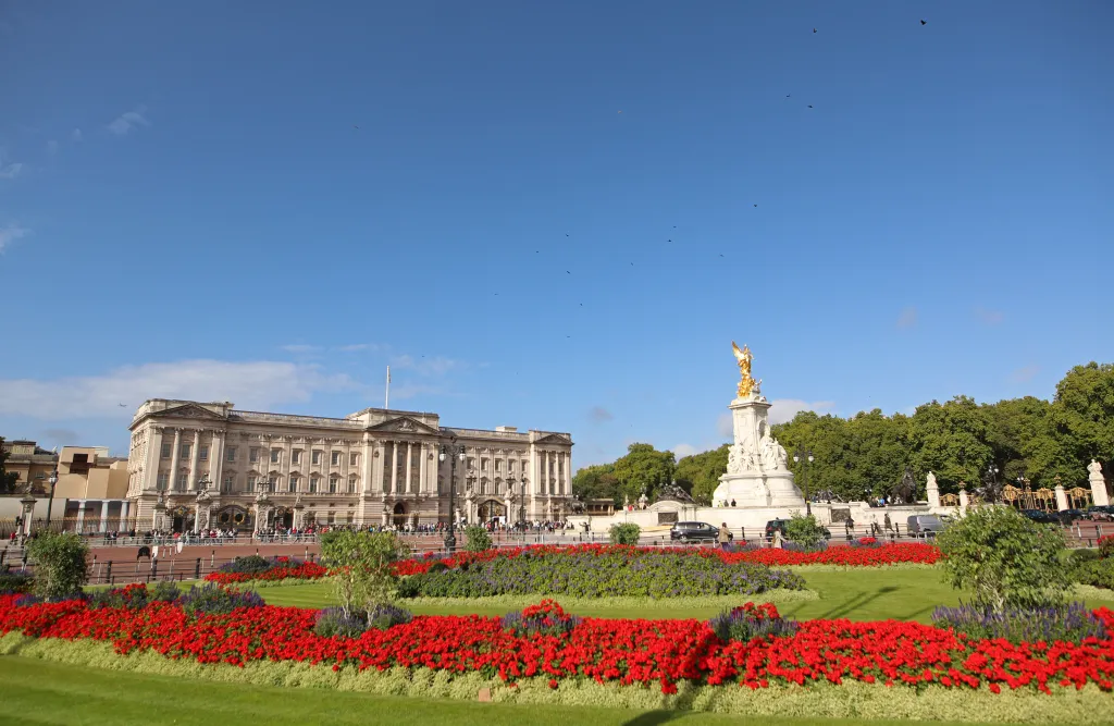 Buckingham Palace with a golden statue in front, surrounded by red flowers and green lawns under a blue sky.