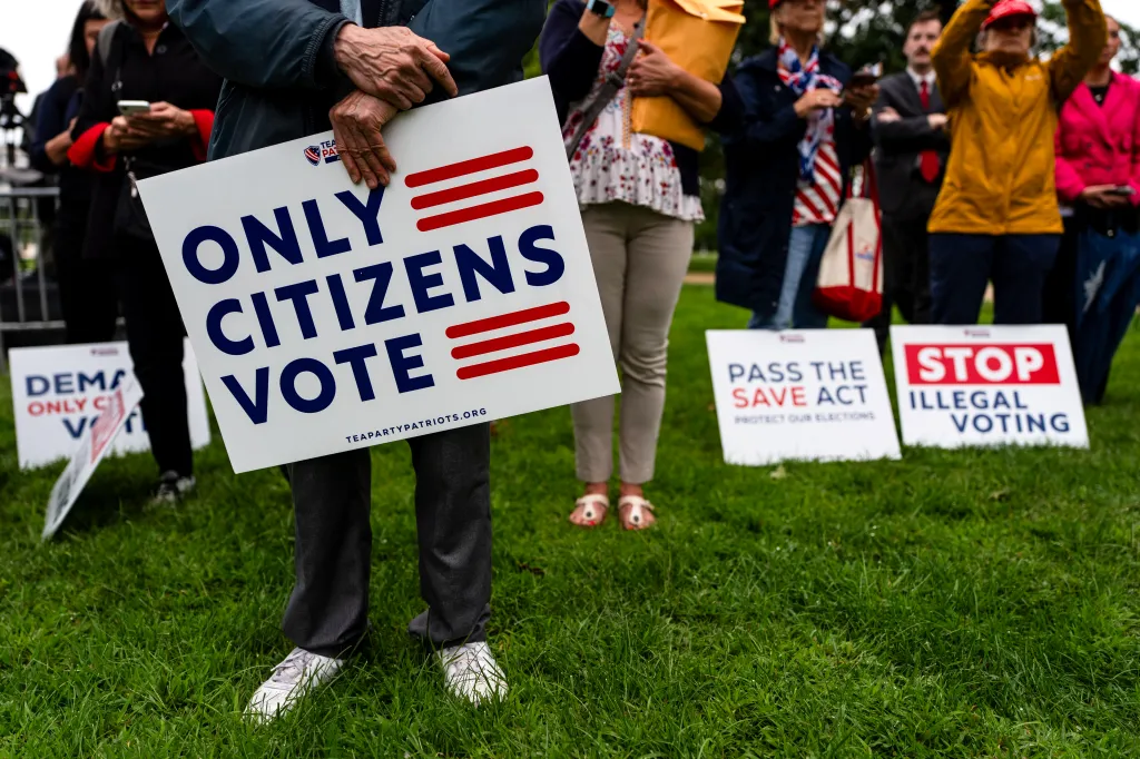 A person holds a sign reading "Only Citizens Vote," while other signs in the background say "Pass the SAVE Act" and "Stop Illegal Voting."