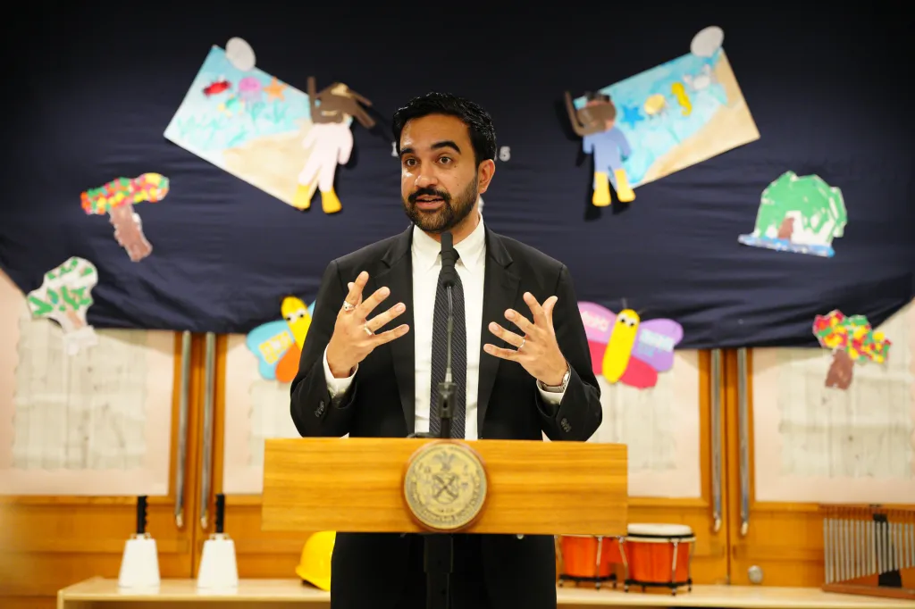 Mayor Zohran Mamdani speaks at a podium in front of a dark blue backdrop adorned with colorful children's art.