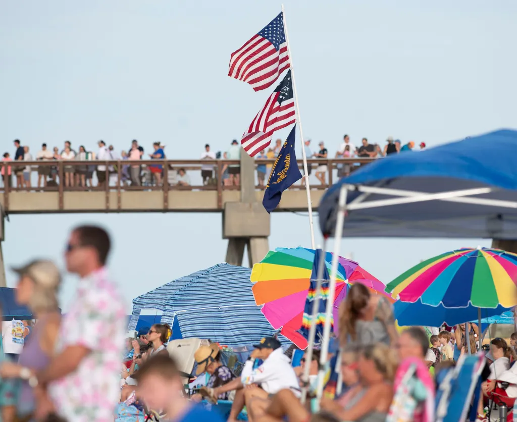 American and Florida state flags fly above a crowd on a beach, with people on a pier in the background.