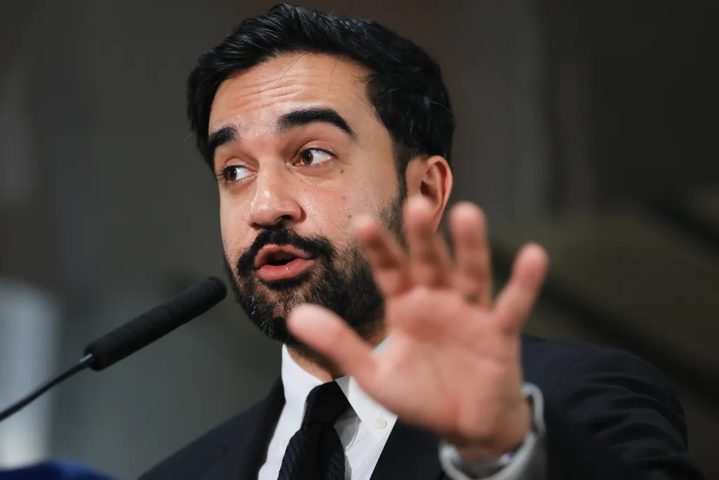 NYC Mayor Zohran Mamdani speaks at a news conference, with his right hand extended forward.