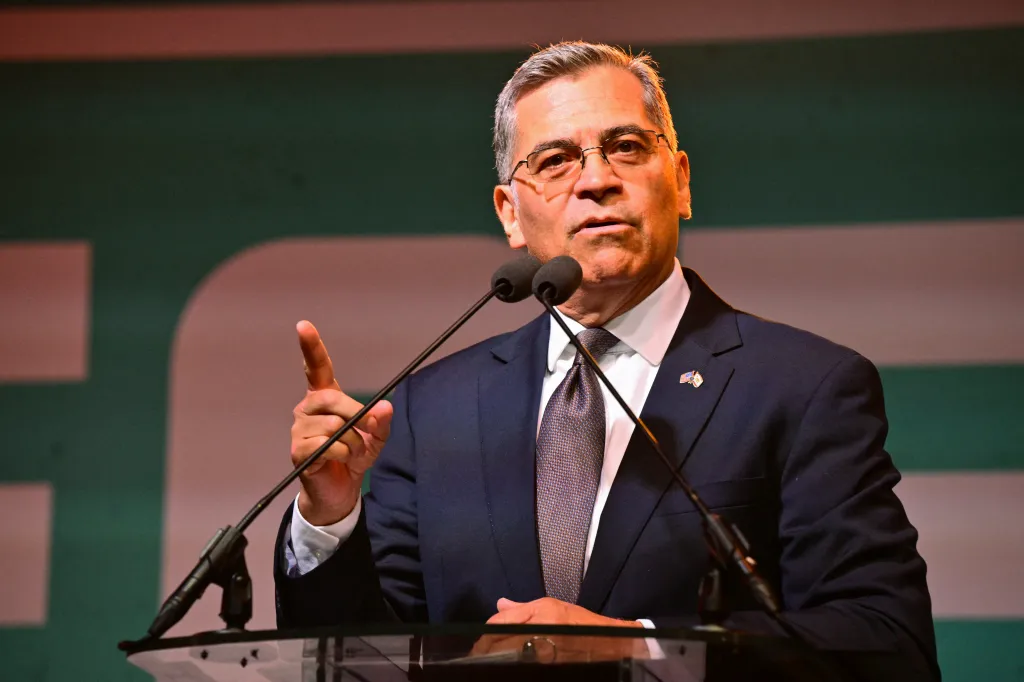 Xavier Becerra speaking at a podium with two microphones, pointing his right index finger upwards.