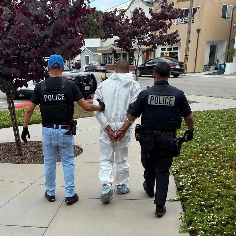 Two police officers arresting a person in a white forensic suit and handcuffs.