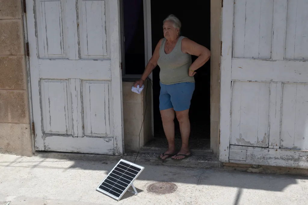 A woman stands in a doorway, holding a white cloth connected by a wire to a small solar panel on the ground.