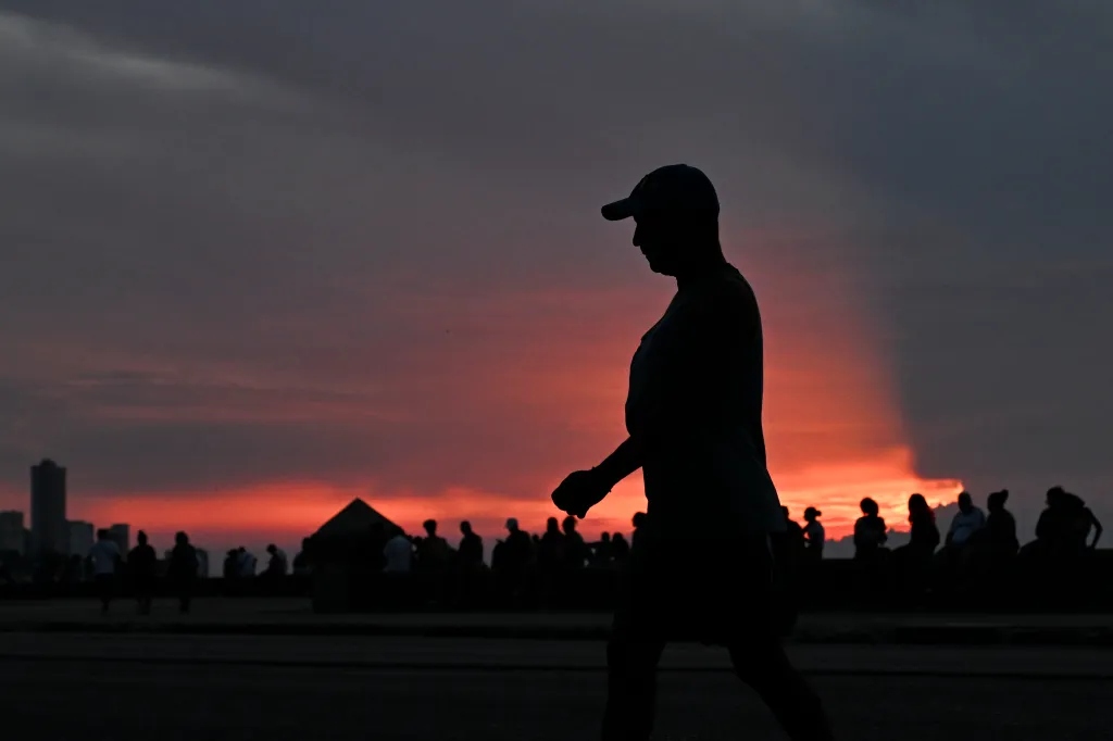 A man in silhouette walks along the Malecon during a nationwide blackout in Havana, with a fiery orange sunset in the background.