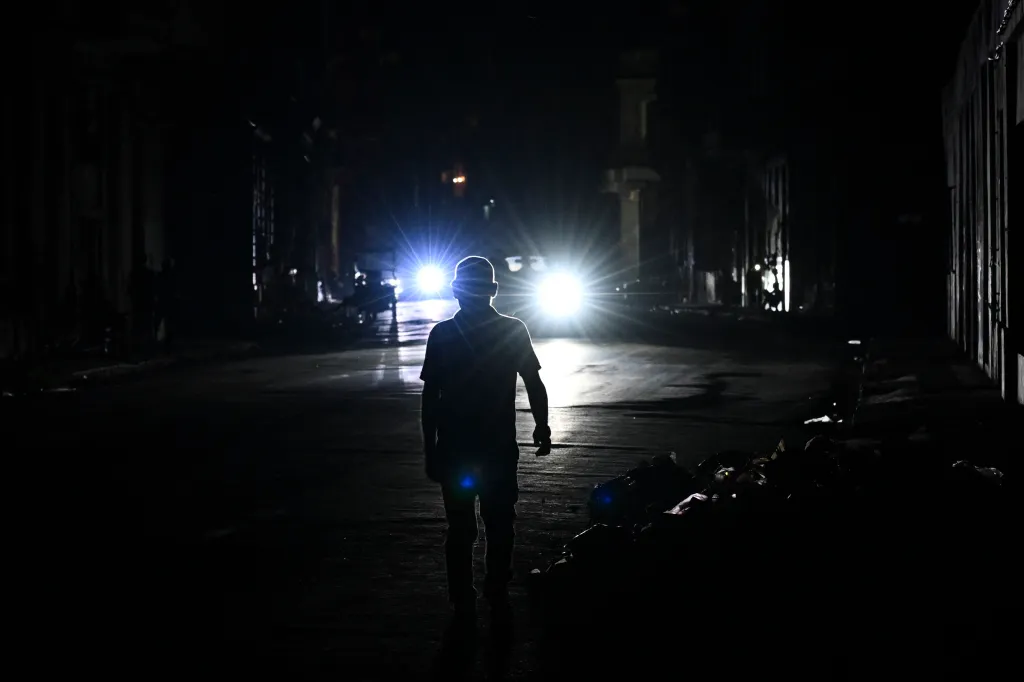 A man walks in silhouette on a dark street, lit from behind by the bright headlights of an approaching vehicle.