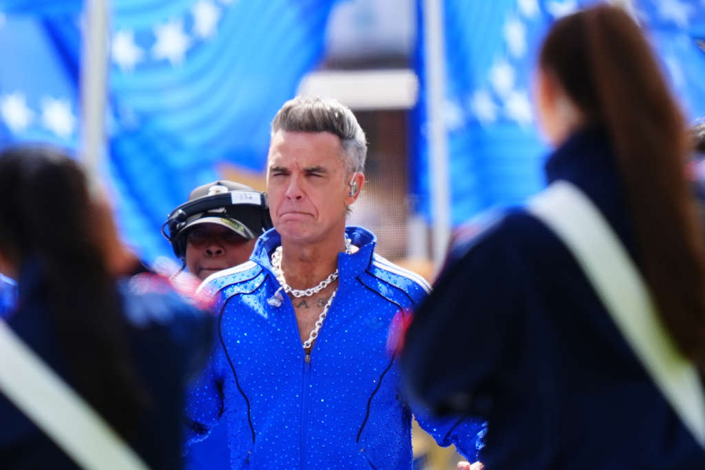 Robbie Williams in a sparkling blue jacket and thick chain necklace, with a slightly pained expression.
