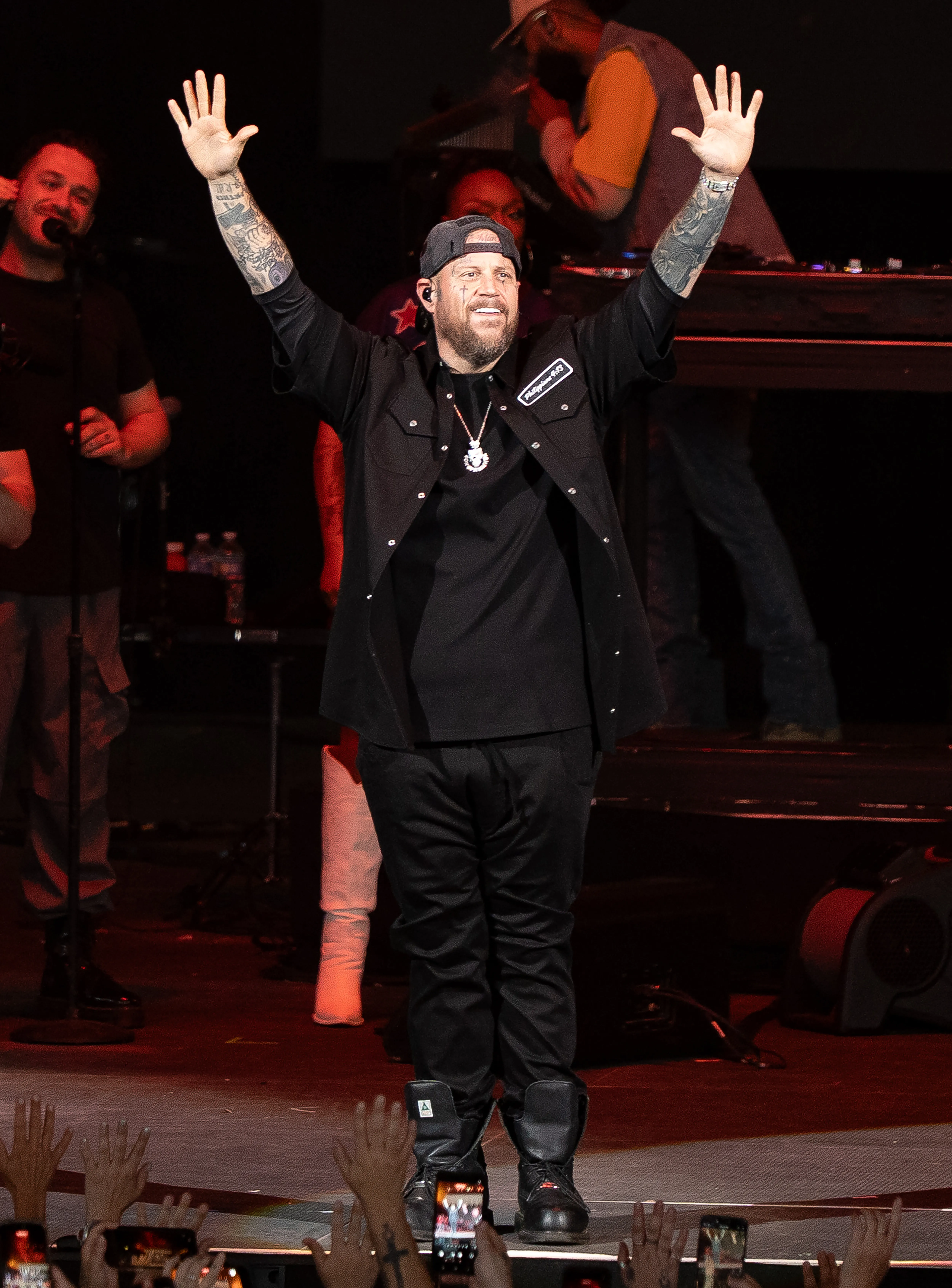 Jelly Roll on stage with both arms raised and tattooed forearms visible.