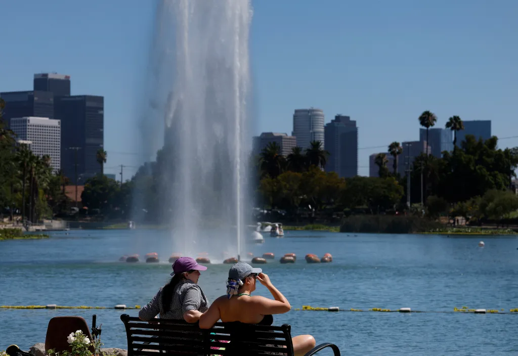 Two people sit on a bench in Echo Park, Los Angeles, with a large fountain and city skyline in the background.