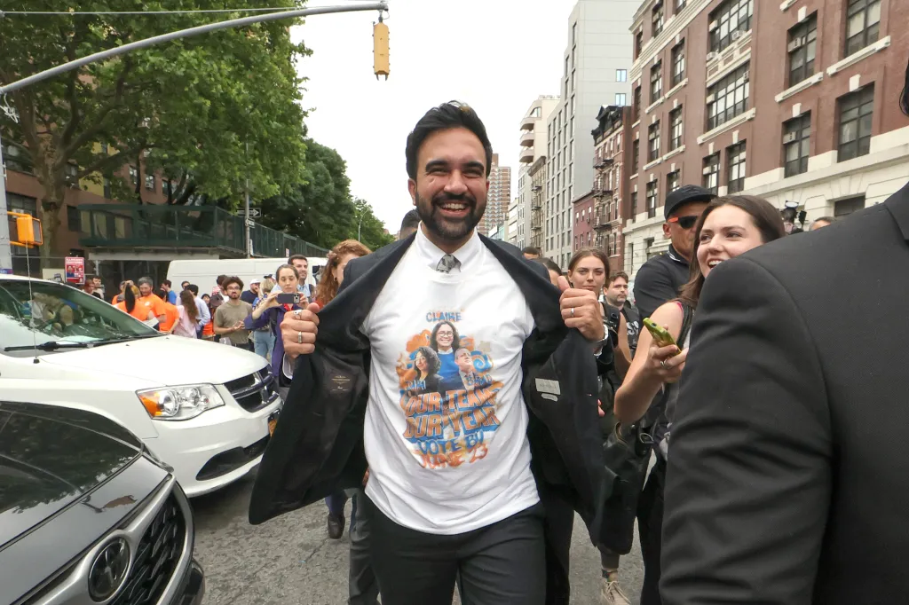 Mayor Zohran Mamdani pulling open his suit jacket to reveal a white t-shirt with images of political candidates.