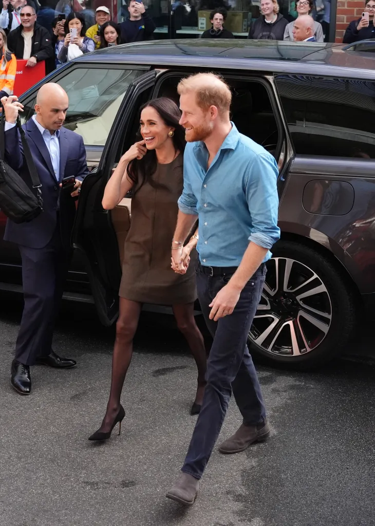 Meghan, Duchess of Sussex, and Prince Harry, Duke of Sussex, holding hands and smiling while exiting a car with people watching in the background.