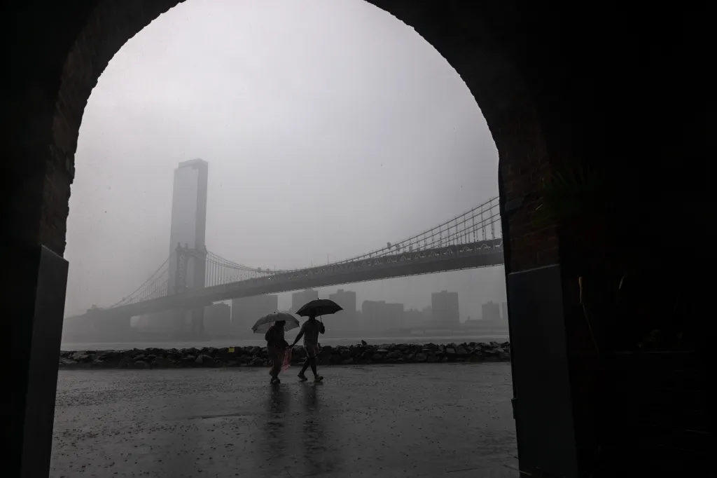 Two people with umbrellas walk in the rain near the Manhattan Bridge and a tall building in Brooklyn.