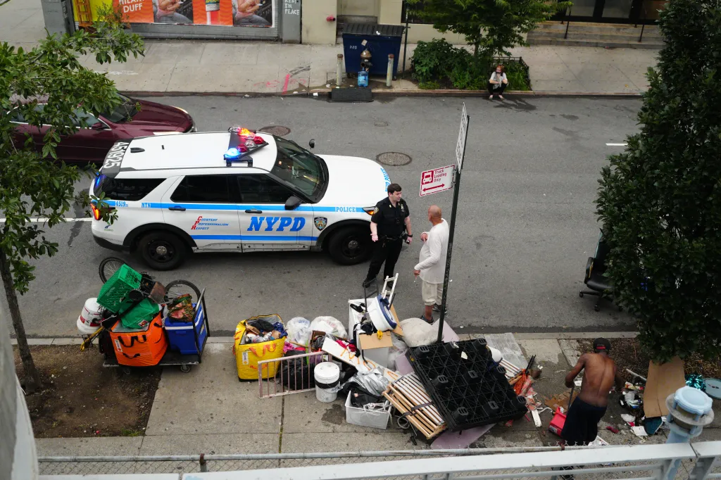 Police officer and civilian next to an NYPD vehicle, with piles of belongings from a homeless encampment.