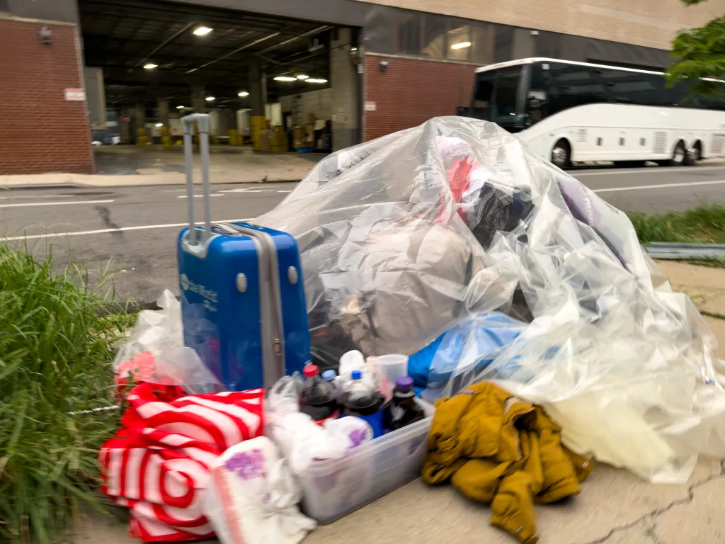 A homeless encampment with a blue suitcase, plastic-covered belongings, and a yellow jacket on a sidewalk.