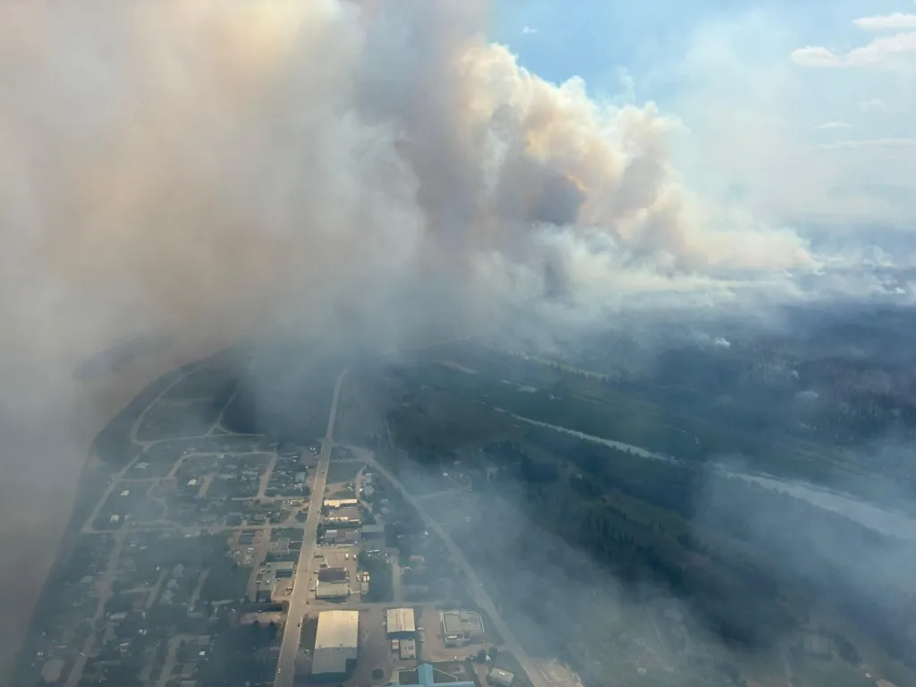 Aerial view of the Fort Simpson wildfire burning out of control, with smoke obscuring parts of the town and surrounding forests.