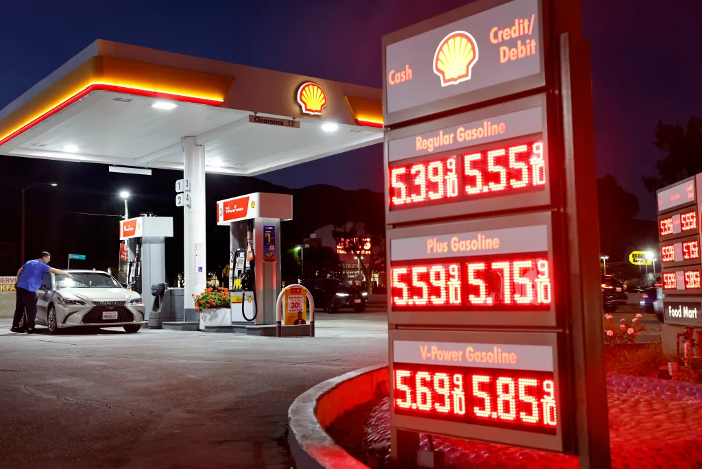 A customer washes their windshield at a Shell gas station where gas prices are displayed at $5.39 for regular, $5.59 for plus, and $5.69 for V-Power gasoline.