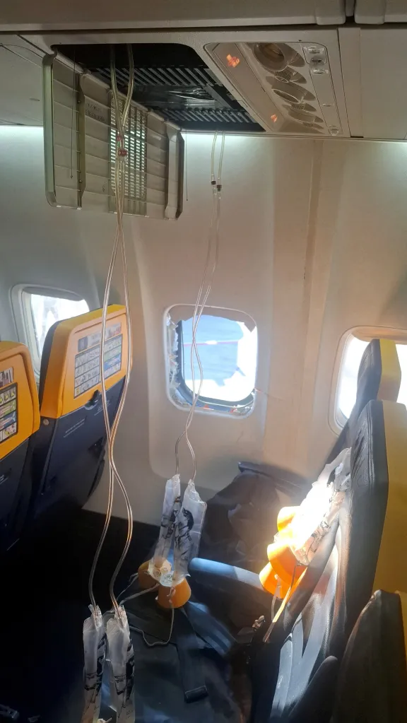 Interior of a Ryanair aircraft showing oxygen masks deployed and a broken window, following an emergency landing in Thessaloniki.