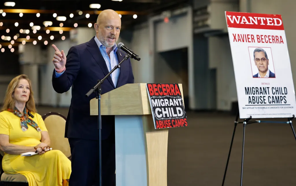 California Gubernatorial candidate Steve Hilton speaks at a news conference, with "Migrant Child Abuse Camps" signs visible.