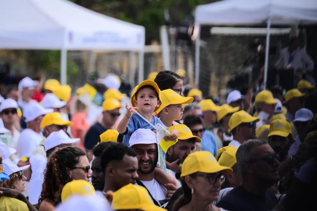 A crowd of people, many wearing white and yellow hats, look forward while a child in a blue shirt on an adult's shoulders points.