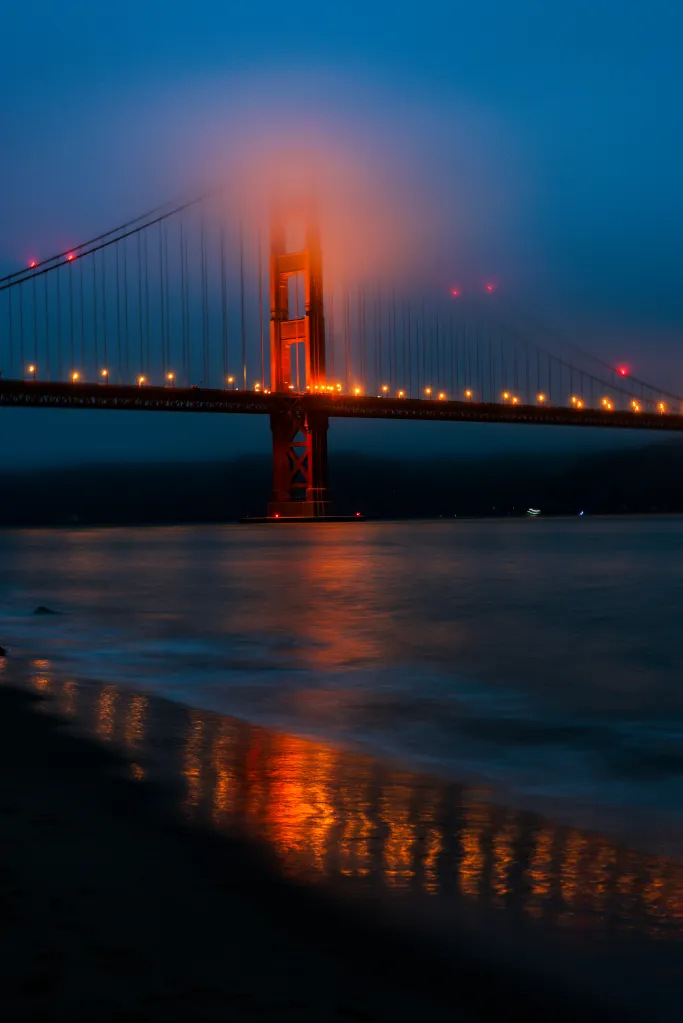 The Golden Gate Bridge at night, partially obscured by fog, with its lights reflecting on the water.