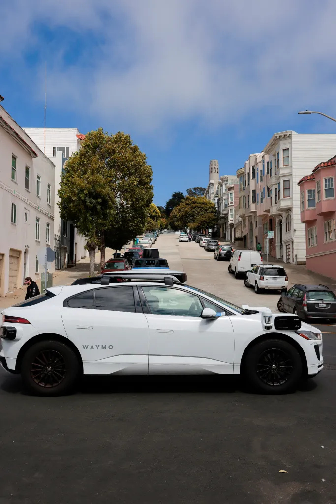 A white Waymo robotaxi with a rooftop sensor unit is parked on a San Francisco street, with Coit Tower visible in the distance.