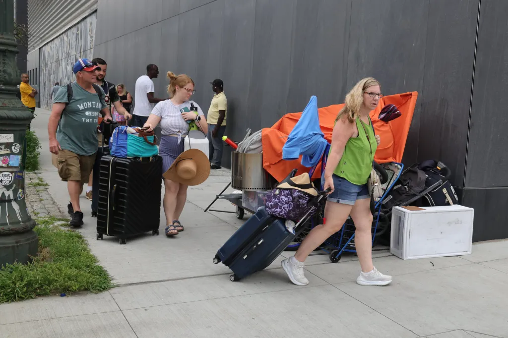 Passengers pulling luggage walk past a homeless encampment with orange and blue tarps along a sidewalk in Midtown Manhattan.