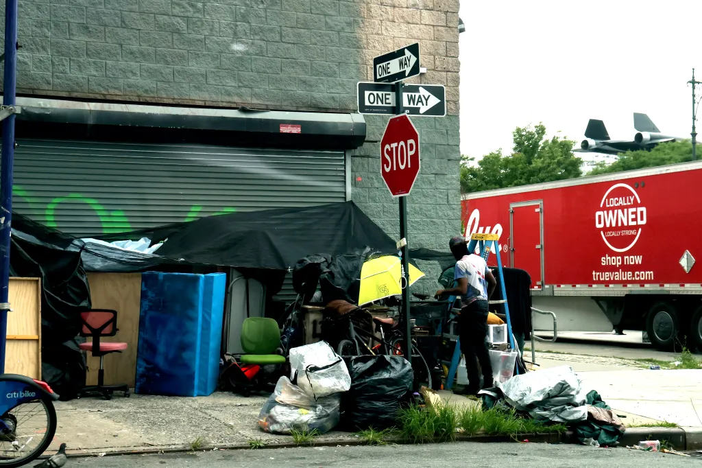 A homeless encampment with a person standing next to a makeshift shelter, with a "One Way" and "Stop" street sign visible, and a red True Value truck and an airplane in the background.