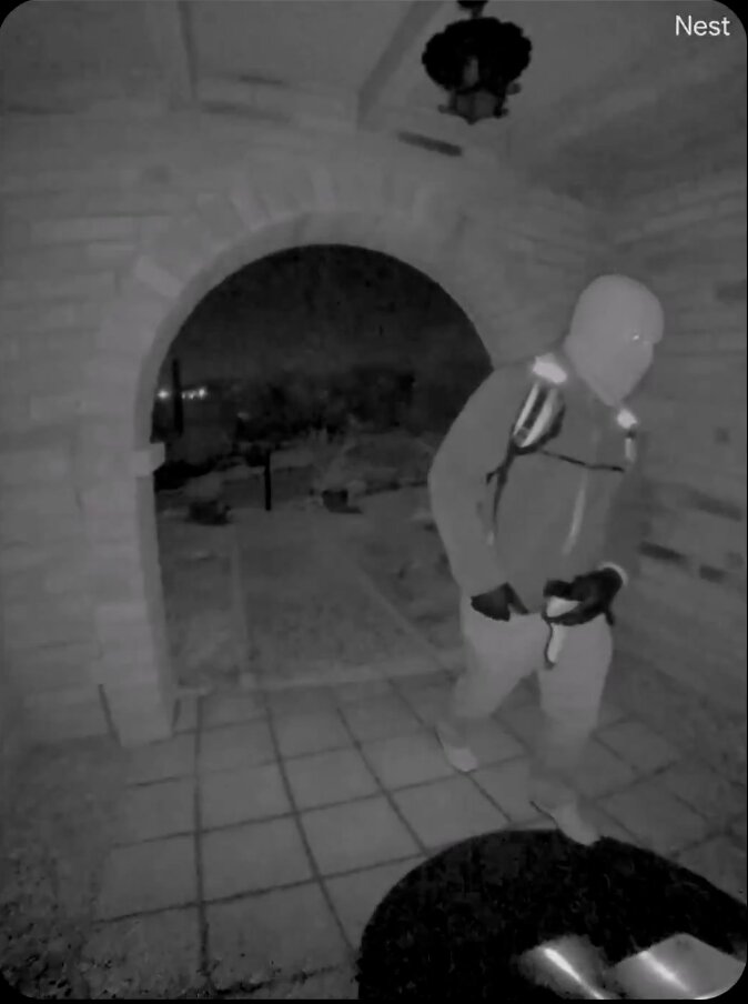 Night vision security footage of a suspect in a balaclava and backpack walking through an arched doorway.