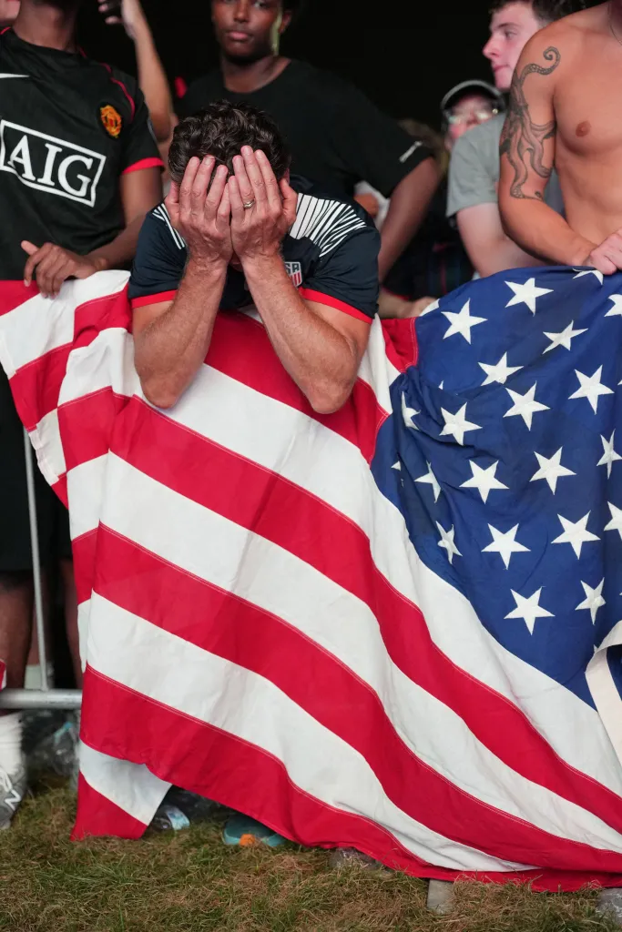 A US soccer fan covers his face in disappointment behind an American flag.