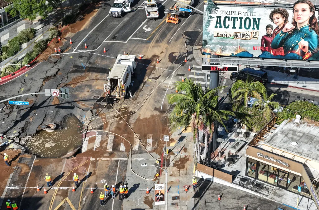 Aerial view of a sinkhole on Sunset Boulevard where a water main break flooded surrounding streets, with utility repair workers present.
