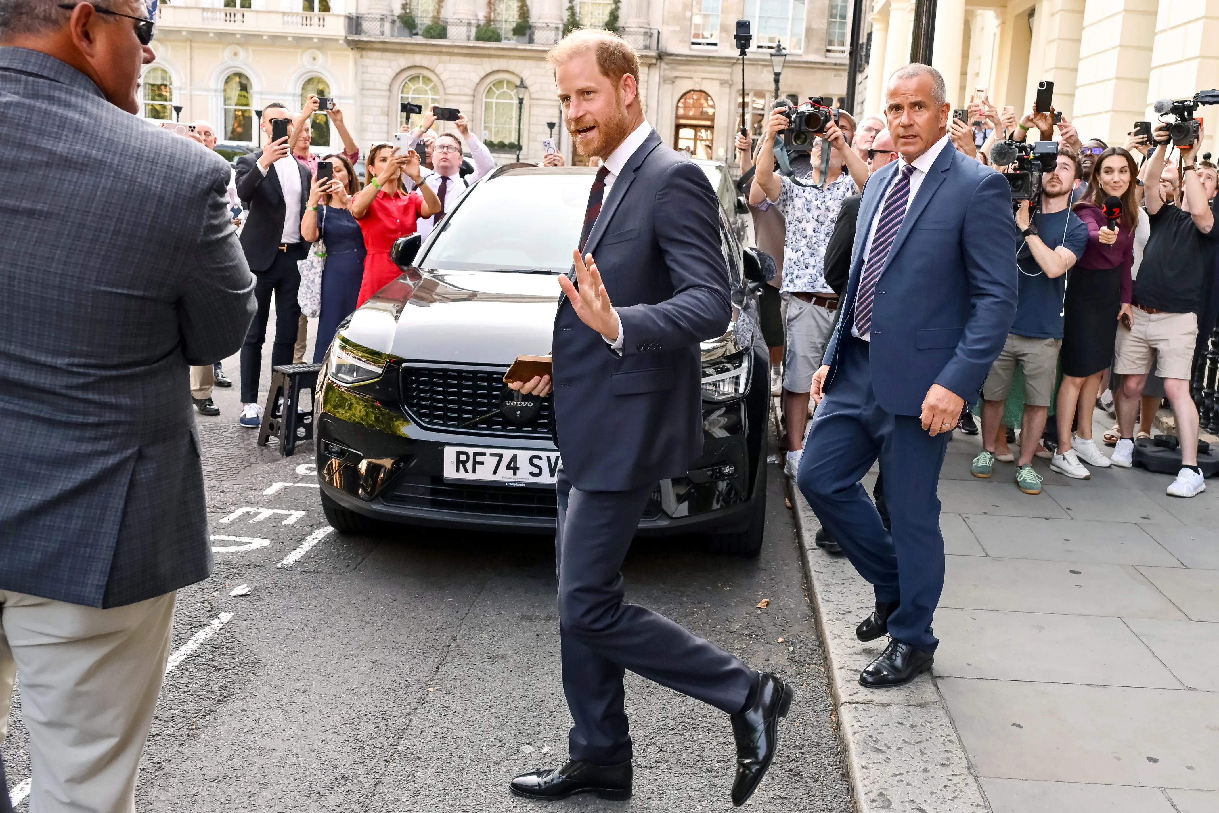 Prince Harry waves as he walks past a crowd of people holding up phones and cameras.