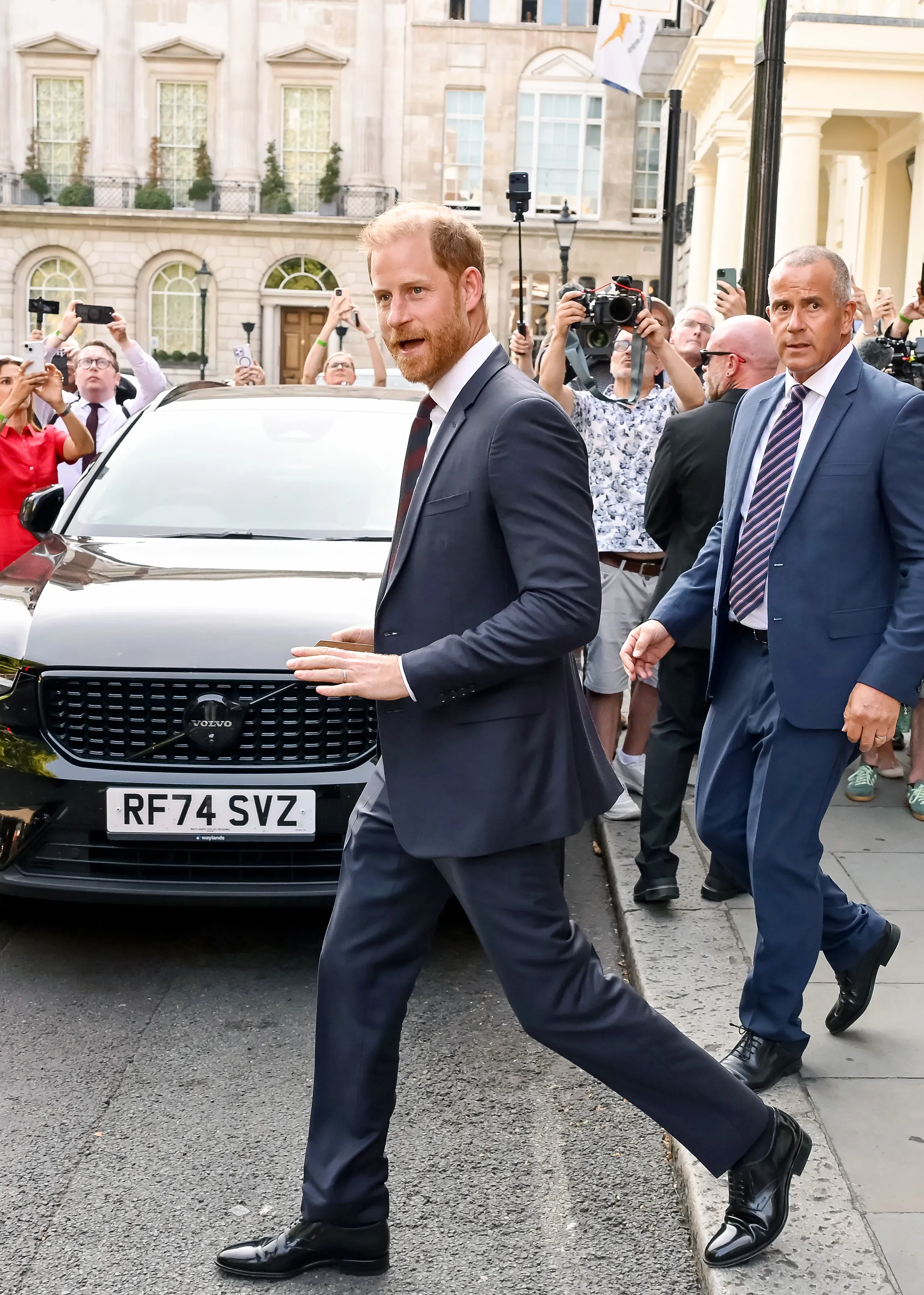 Prince Harry, Duke of Sussex, walks past a black Volvo with a security guard next to him, while people take photos in the background.
