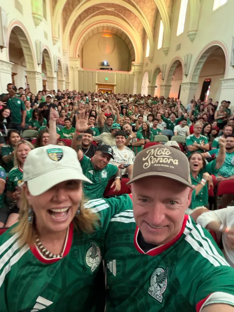 Senator Mark Kelly and Gabby Giffords wearing Mexico jerseys, posing for a selfie in a large hall filled with cheering soccer fans.