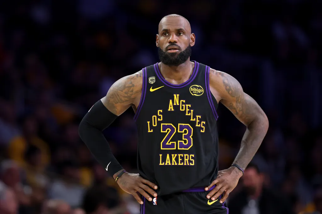 LeBron James in a black Los Angeles Lakers jersey with "23" in yellow and purple, looking on during a game.