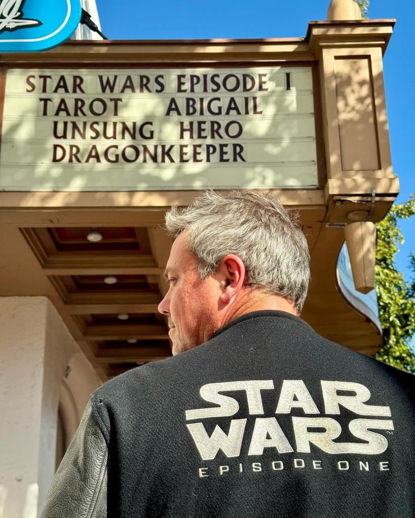 A man in a "Star Wars Episode One" jacket looks up at a movie marquee advertising "Star Wars Episode I".