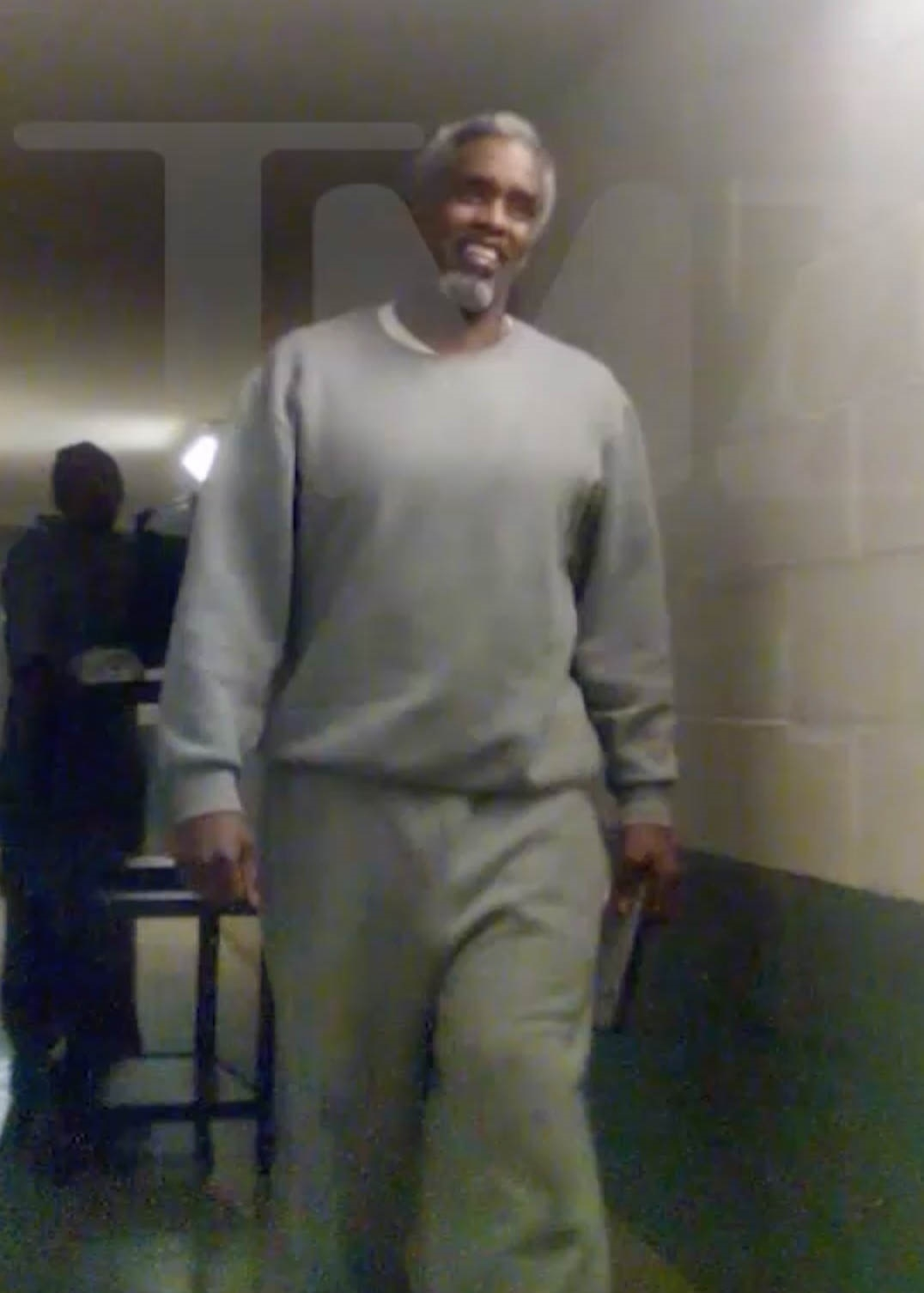 Diddy, Sean Combs, smiling in a grey prison uniform with gray hair and a beard, walks down a hallway with another inmate in the background.