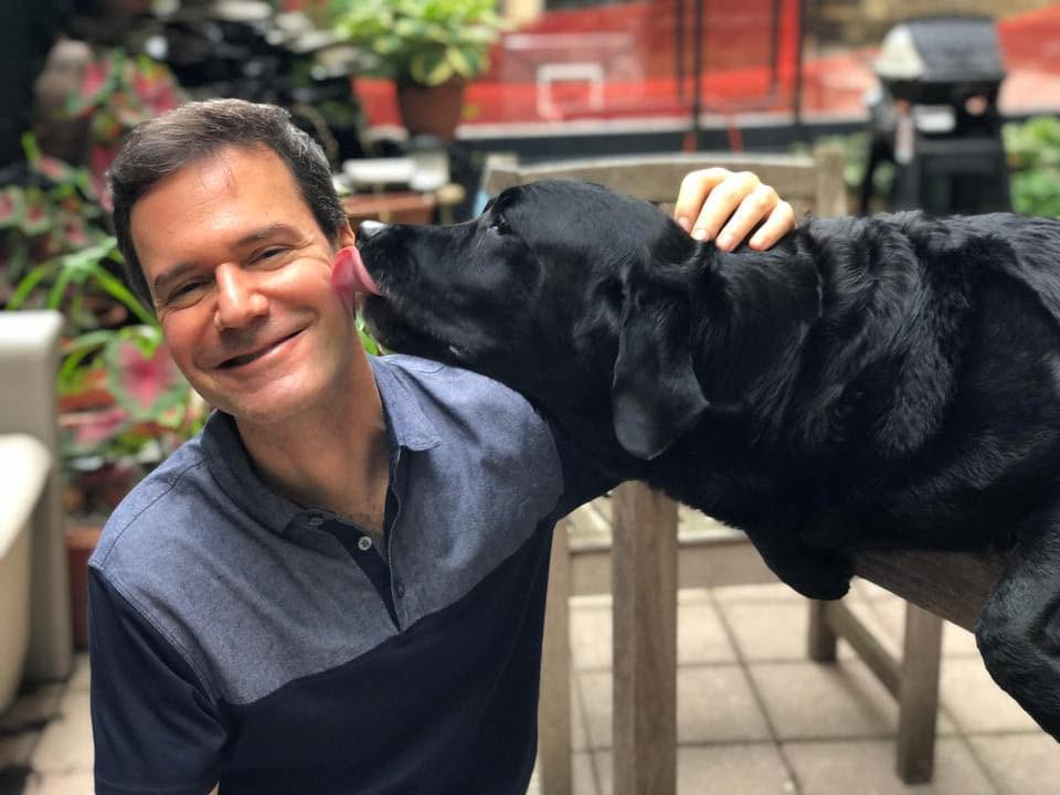 Moshe Grunhaus smiling as a black dog licks his ear.