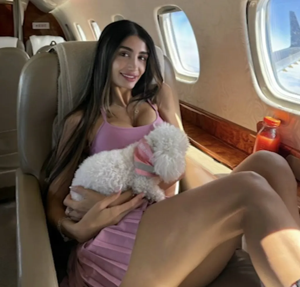 Adva Lavie, also known as Mia Ventura, seated inside a private jet, holding a small white dog with a pink "SERVICE" vest.