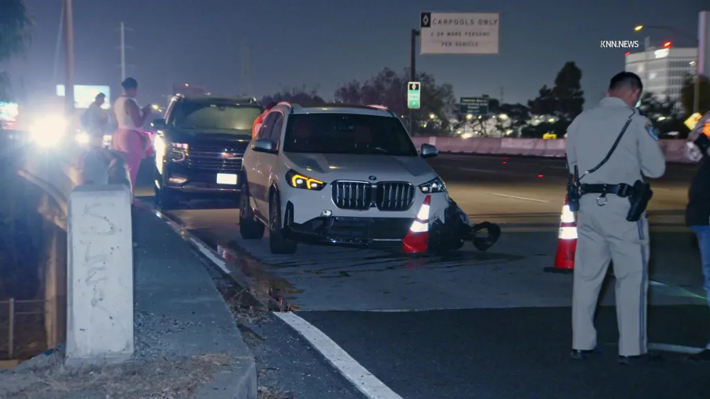 A white BMW and a dark SUV are stopped on a freeway at night following a multi-vehicle crash, with a police officer and others standing nearby.