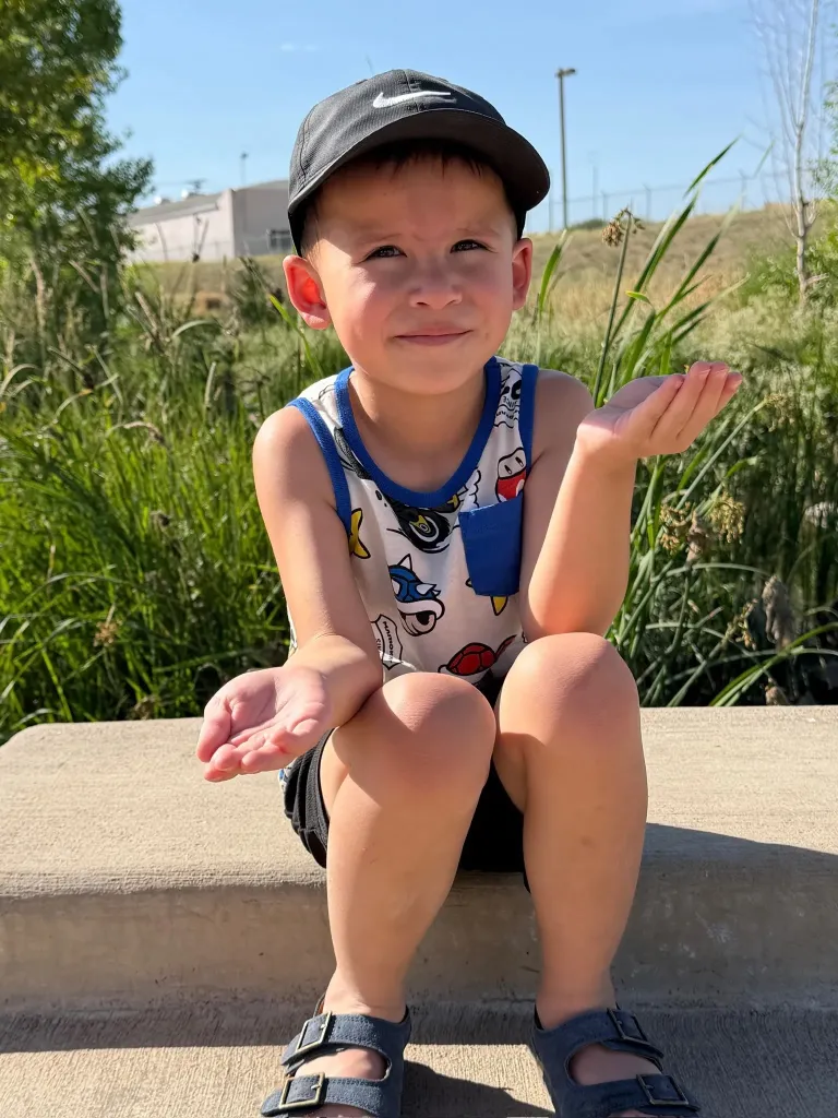 A young boy wearing a cap and tank top, sitting with his palms up.