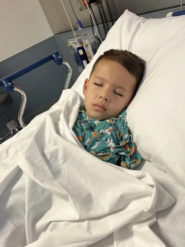 A young boy with closed eyes rests in a hospital bed, wearing a green patterned shirt.