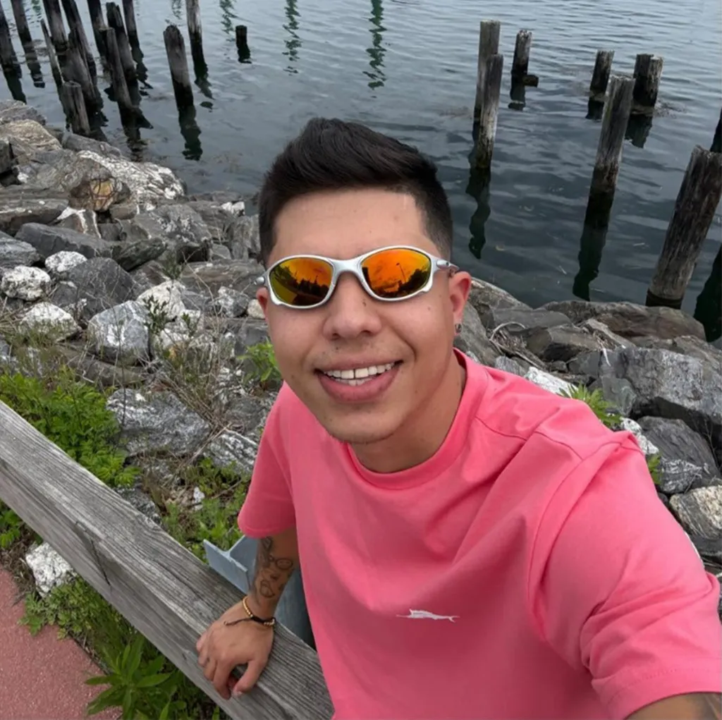 Joan Sebastian Guerrero smiling in a pink shirt and sunglasses, next to water with wooden posts.