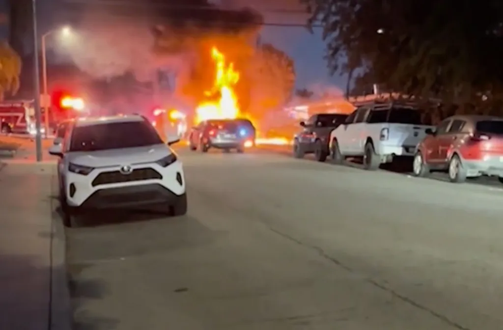 A street at dusk with a car fully engulfed in flames, parked among other vehicles, with a firetruck in the background.