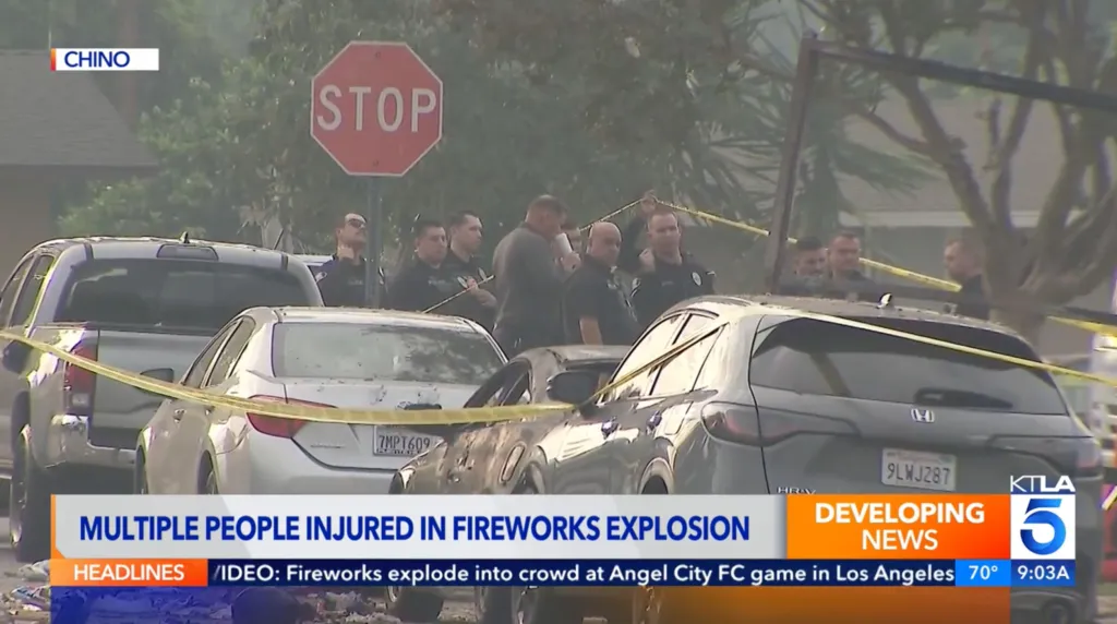 Police stand behind yellow caution tape at a fireworks explosion site in Chino, with two damaged cars in the foreground.