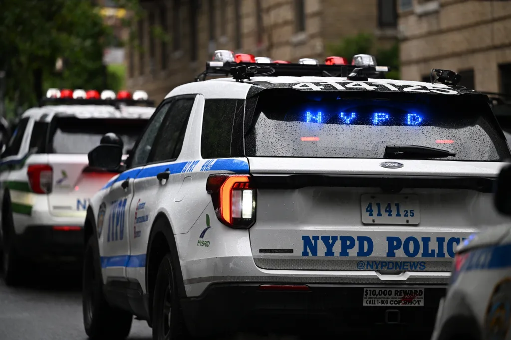 Two NYPD patrol cars, one with "NYPD" illuminated in blue on its rear window, parked on a street.