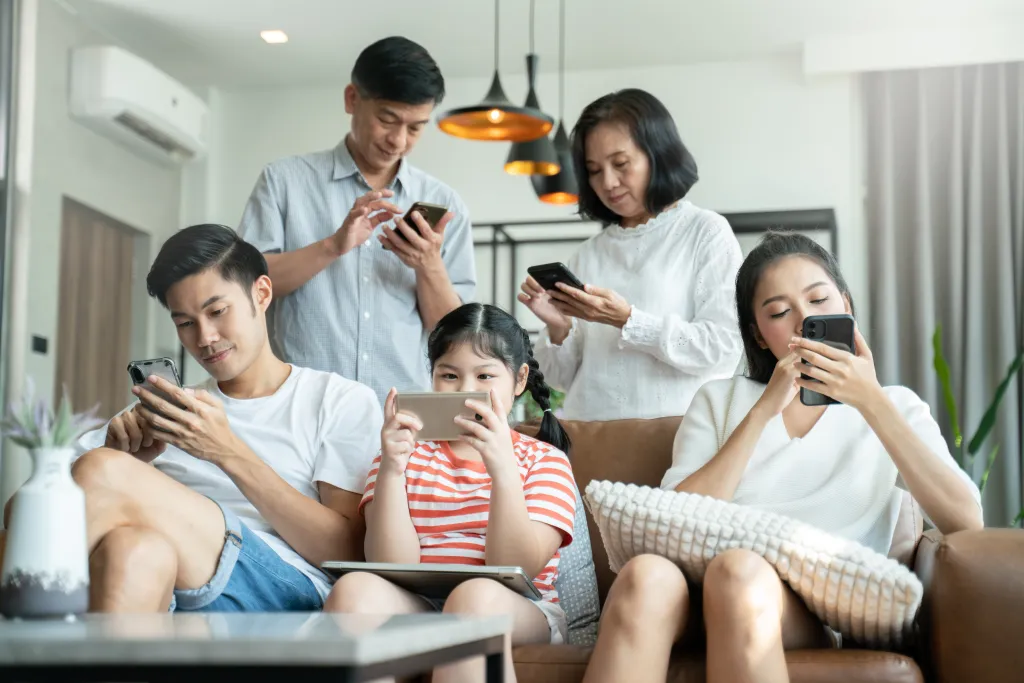 An Asian family of five sitting in a living room, each looking at a smartphone or tablet.