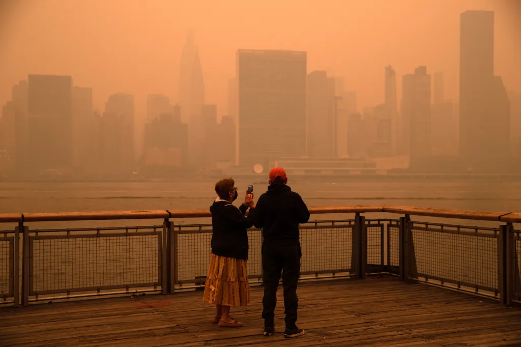 Two people on a pier photograph the Manhattan skyline obscured by orange wildfire haze.